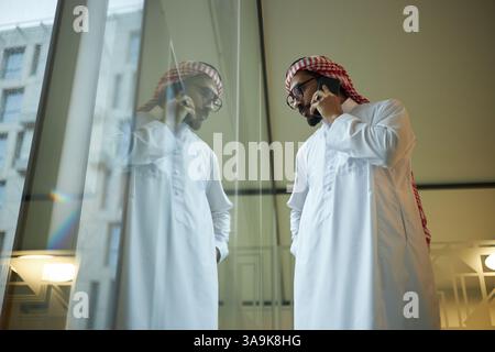 Low angle view medium long shot of young Middle Eastern businessman standing in front window, talking on phone Stock Photo