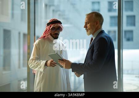 Young Middle Eastern and mature Caucasian businessmen standing in office, checking data on digital tablet Stock Photo