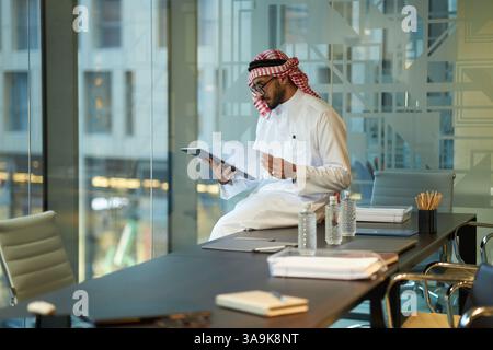 Serious young Middle Eastern businessman in traditional clothes sitting on table in board room working with important documents Stock Photo