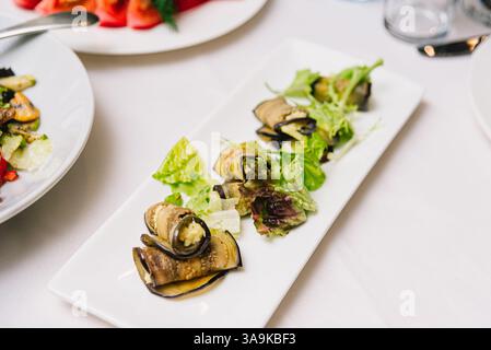 Appetizer of eggplant slices rolled and filled with cheese or vegetables, served with fresh lettuce on a rectangular white plate. Stock Photo