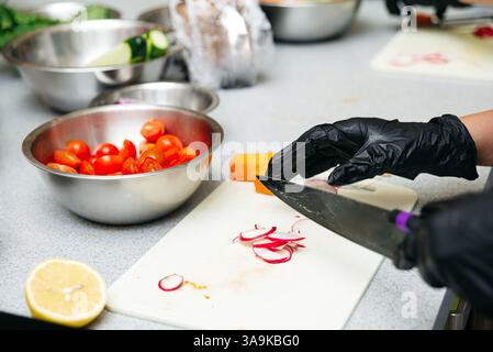 Close-up of a gloved hand slicing radishes on a cutting board with tomatoes, cucumbers, and lemon prepared for salad. Stock Photo