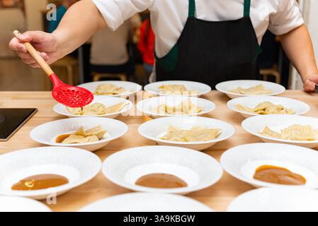 A chef in a black apron plates freshly cooked ravioli with sauce into multiple white dishes, preparing for serving in a restaurant. Stock Photo