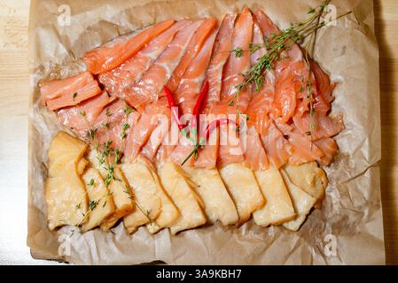 Assorted sliced smoked and cured fish, garnished with red chili peppers and fresh thyme on parchment-lined wooden tray. Stock Photo