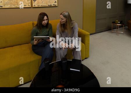 Discussion between two women in a modern lounge area while using digital devices and experiencing professional collaboration Stock Photo