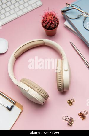Modern workspace featuring wireless headphones, keyboard, mouse, stationery, glasses, and a cactus on a pink background Stock Photo