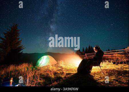 Hiker woman relaxes by campfire, illuminated by its warm glow, with tent nearby under stunning, star-filled sky. Milky Way stretches overhead, adding magical touch to serene mountain setting. Stock Photo