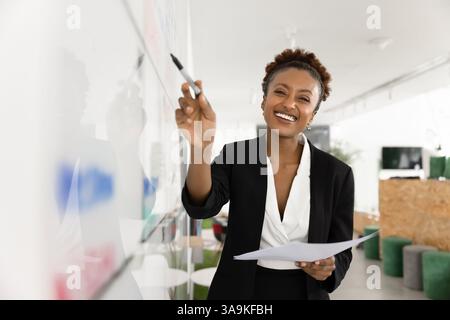 Cheerful positive young Black businesswoman writing on whiteboard Stock Photo