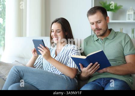 Couple reading paper book and ebbok sitting on a couch at home Stock Photo