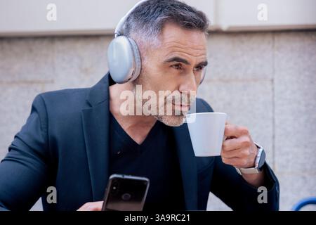 Mature caucasian businessman with headphones drinking coffee and using smartphone Stock Photo