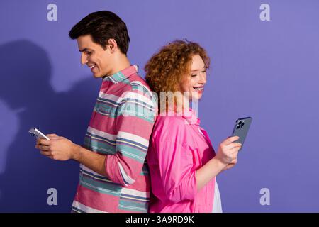 Young man and woman standing back to back using smartphones against vibrant purple background showcasing casual fashion Stock Photo