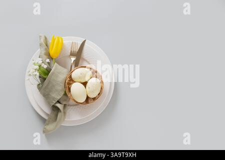 Easter table setting with spring yellow tulip, white plates, bunny, eggs in nest isolated on gray background. Happy Easter celebration. Top view. Copy Stock Photo