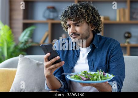 A young man with curly hair checks his smartphone while holding a plate of salad at home on the couch during lunchtime. Stock Photo