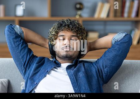 Young man resting and relaxing at home, lying on the couch with headphones, listening to music. Guy close up in the living room with his hands behind his head thinking and dozing. Stock Photo