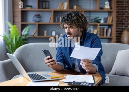 A young man looks concerned while reviewing a bill and using a mobile phone while sitting at his desk with a laptop at home. Stock Photo