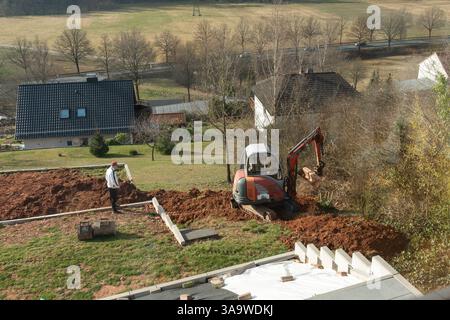 Excavator actively digging in a residential construction site with a worker supervising the process. Houses surrounded by fields and trees in the Stock Photo