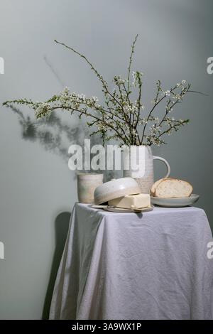 Minimal rustic breakfast setup with ceramic pitcher containing blooming cherry branches butter dish bread slice on plate ceramic cup on linen tableclo Stock Photo