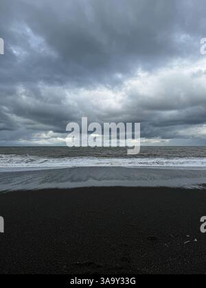 Dark clouds over ocean waves rolling onto a black sand beach, creating a moody and dramatic atmosphere. Stock Photo