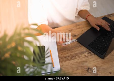 Portrait of smiling senior freelance businesswoman working from home, using laptop computer and smartphone sitting at table, typing, chatting online. Stock Photo