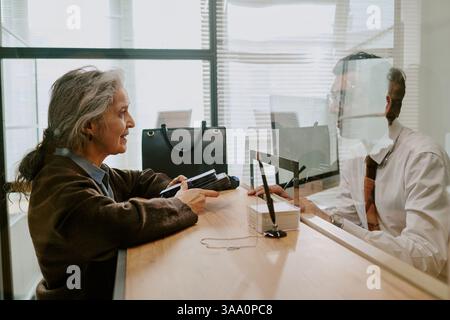 Senior woman having conversation with young man through glass partition at office desk, holding documents and sitting comfortably, highlighting importance of communication in business settings Stock Photo