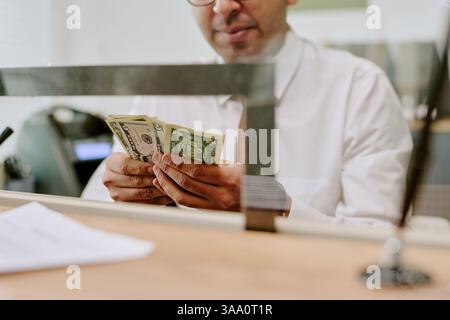 Bank employee counting cash bills at counter behind transparent glass partition in workplace setting in financial institution holding currency in hands Stock Photo