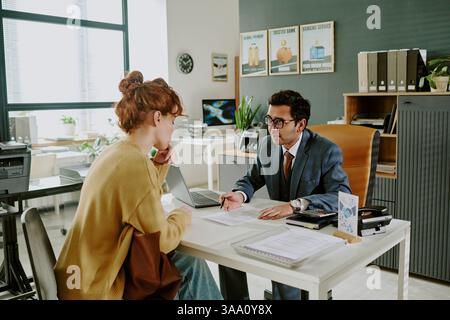 Business meeting scene depicting two professionals engaging in discussion about finances while sitting at office desk with documents and devices spread out around them Stock Photo