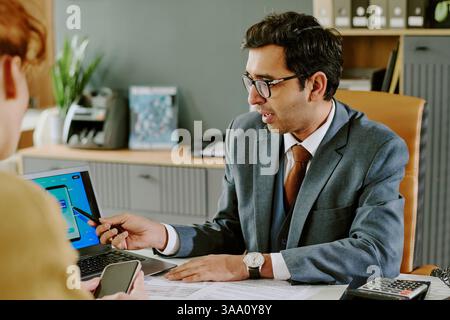 Indian, businessman in formal suit presenting information on digital tablet in modern office setting with wooden decor, engaging with colleague for work-related discussion Stock Photo