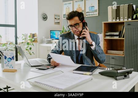 Portrait of dedicated businessman talking on phone while reviewing documents at modern office desk filled with technology and office supplies Stock Photo