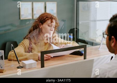 Young woman with curly hair sitting at reception desk while handling paperwork, interacting with unseen individual, including various documents and office supplies for transaction Stock Photo