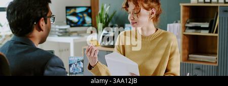 Colleagues in modern office kindly exchanging business cards while discussing project details, with various documents and office supplies visible in background Stock Photo