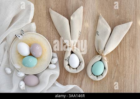 Easter eggs in pastel colors on linen cloth on a light wooden table. Easter eggs wrapped in a napkins folded like bunny ears. Happy Easter concept. To Stock Photo