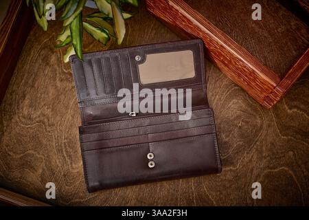 Open brown leather wallet displayed on a wooden table beside a leafy plant. Features card slots, a clear ID window, and a zippered pocket, offering a Stock Photo