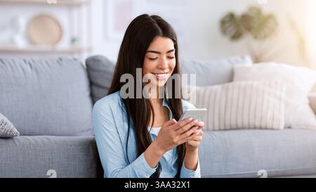 Joyful Korean Girl Texting On Smartphone While Resting At Home Stock Photo