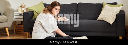 A young woman with Down syndrome sits at home, focused on a creative task in cozy decor. Stock Photo