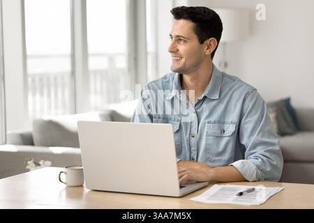 Young man sitting at desk with laptop, smiling, looking away Stock Photo