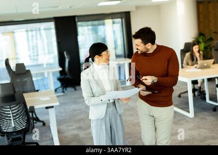 Two professionals engage in a lively discussion while walking through a contemporary office, exchanging documents and ideas. Stock Photo