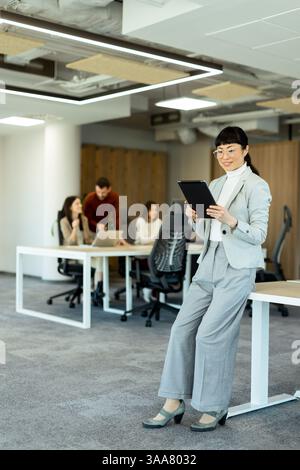 In a modern office, a focused Japanese businesswoman in a stylish suit uses her tablet while team members collaborate in the background, fostering a p Stock Photo