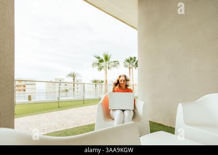 Middle-aged woman in casual outfit sitting on a modern hotel terrace, multitasking with laptop and phone while enjoying a peaceful vacation setting wi Stock Photo