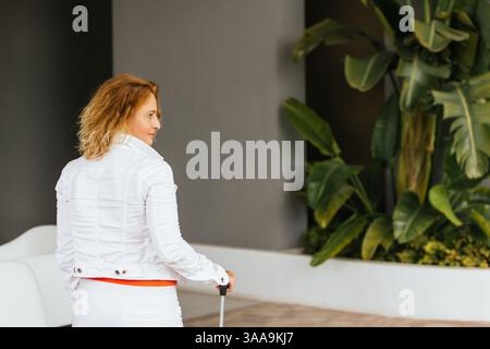 Middle-aged woman in white jacket and pants walking into a hotel pulling a suitcase, seen from behind, with tropical plants and modern decor in the ba Stock Photo