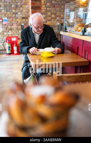 An elderly man sitting at a small table with a cup of coffee reading a book. Stock Photo