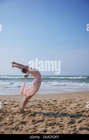 A female dancer in a soft pink dress performs an expressive back arch on a sandy beach. Waves crash behind her as she moves barefoot under the bright blue sky. Stock Photo