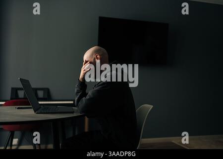 Stressed bald freelancer covering face with hand while sitting at desk in home office, feeling tired and overwhelmed by work. Stock Photo