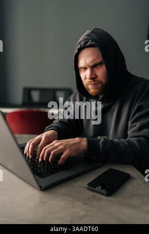 Vertical portrait of hacker male in black hoodie serious looking at camera while typing on laptop keyboard, trying to break password. Concept of Stock Photo
