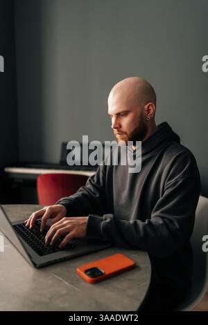 Vertical shot of serious male developer with beard and hoodie remote working from home office sitting at desk and typing on laptop, concentrating on Stock Photo