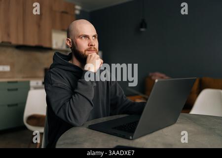 Portrait of pensive freelancer male sitting at table, working on laptop computer while resting hand on chin, deep in thought looking away Stock Photo
