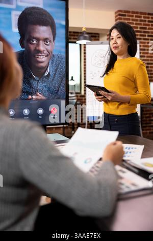 Black businessman on digital monitor, listening to company updates presented by his female colleagues during virtual meeting. Diverse startup team on videoconference, collaborate in brick wall office. Stock Photo
