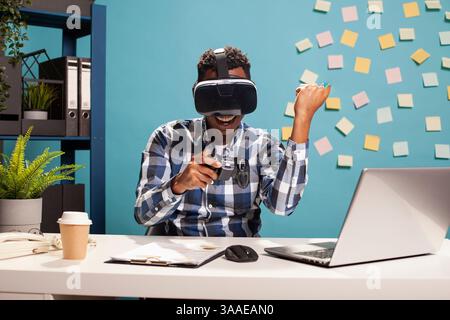 Cheerful male entrepreneur wearing VR goggles, celebrating with his fist raised and game controller in hand. Manager engaged in virtual reality with headset, interacting with a 3D gaming simulation. Stock Photo