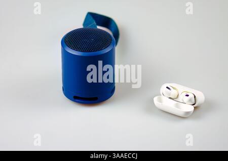 Compact blue speaker with mesh grille and strap next to white wireless earbuds in an open case with purple accents on a plain surface. Stock Photo
