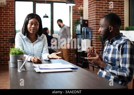 Young black woman explaining her work experiences and expectations to professional hiring manager. Male recruiter and female candidate engage in a formal discussion in a modern office. Stock Photo