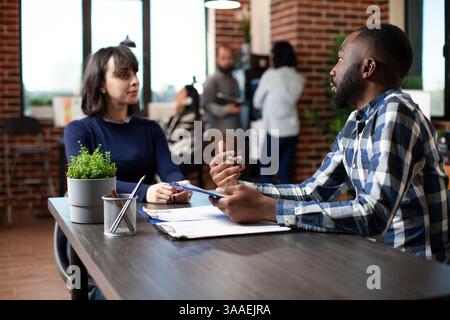 Young female job candidate listening attentively to african american office worker, discussing recruitment process at startup office. HR male representative with clipboard, interviewing a white woman. Stock Photo