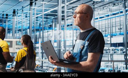 Smart factory senior engineer uses laptop to oversee operation of robotic arms and conveyor systems. Experienced industry 4.0 plant technician monitors system performance using notebook, camera A Stock Photo